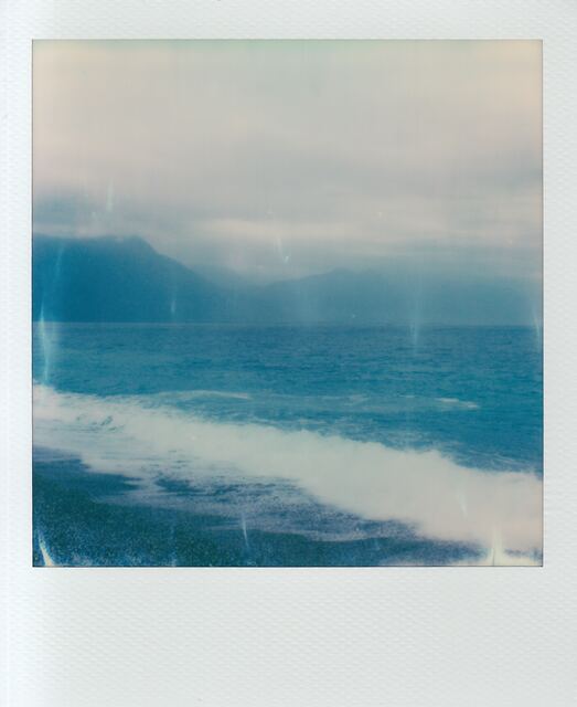 polaroid: a wave breaking on dark sand, mountains in haze behind